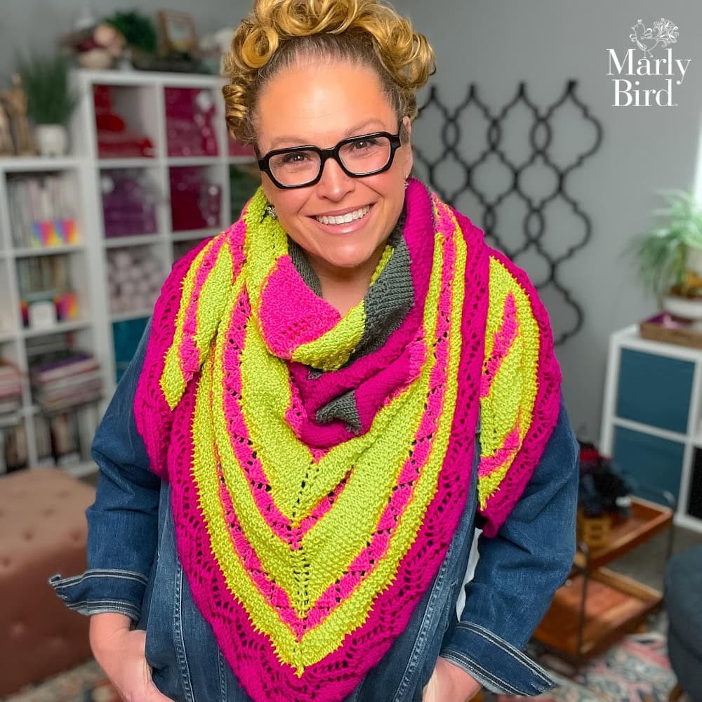 Person models a vibrant knit shawl in pink, yellow, and gray worsted yarn over denim; bookshelves and yarn in background.
