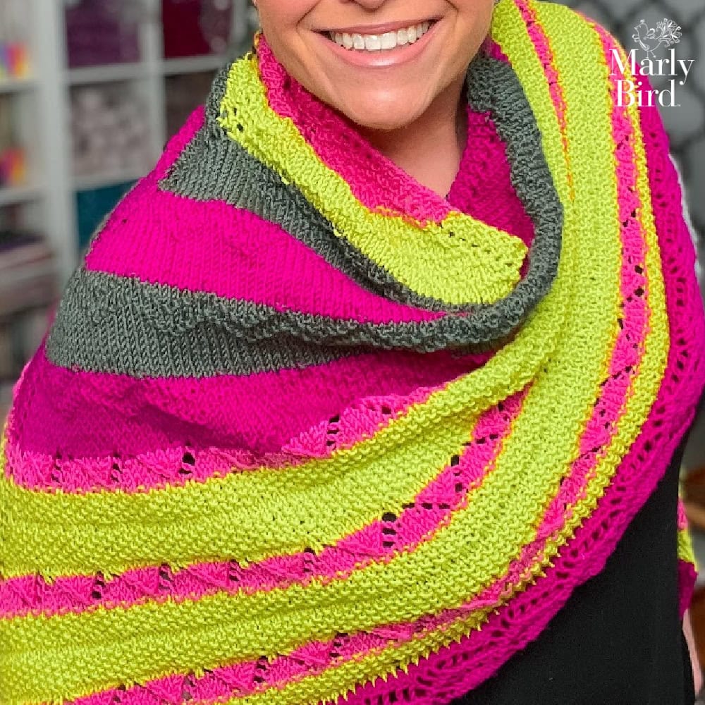 Smiling person models a knitted triangle shawl with bright pink, lime green, and gray stripes; colorful yarn shelves behind.
