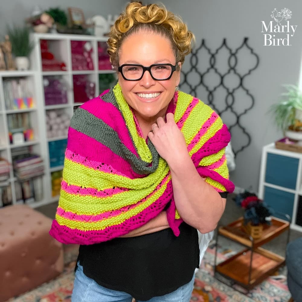 Person models a bright pink, green, and gray knit shawl in a yarn-filled room; "Marly Bird" logo at top right.