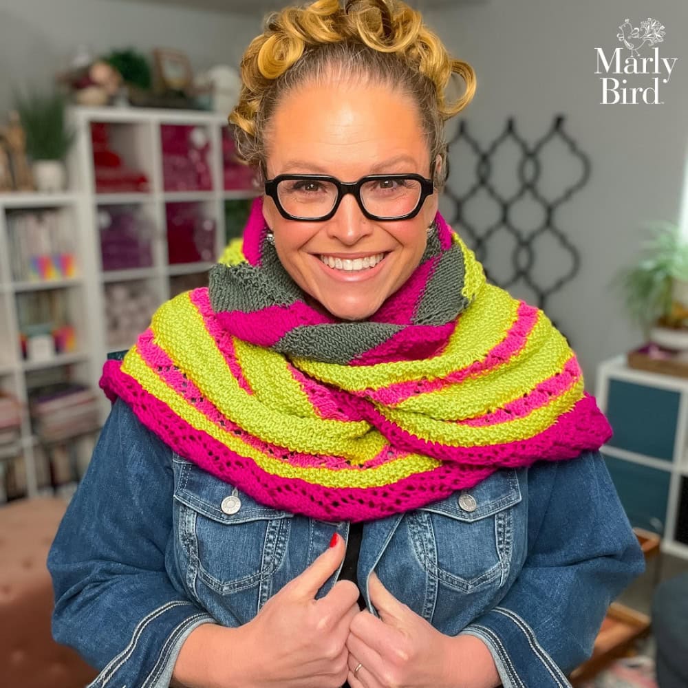 Woman models a knit shawl with pink, lime green, and gray stripes; textured stitches; “Marly Bird” logo in corner.