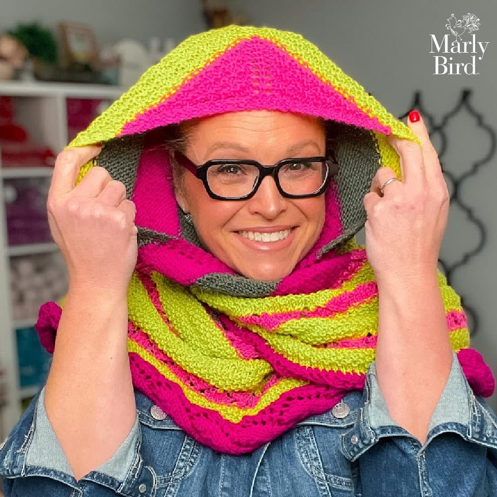 Person wearing a pink and yellow knitted hooded cowl, holding up the hood; yarn shelves in background, Marly Bird logo visible.