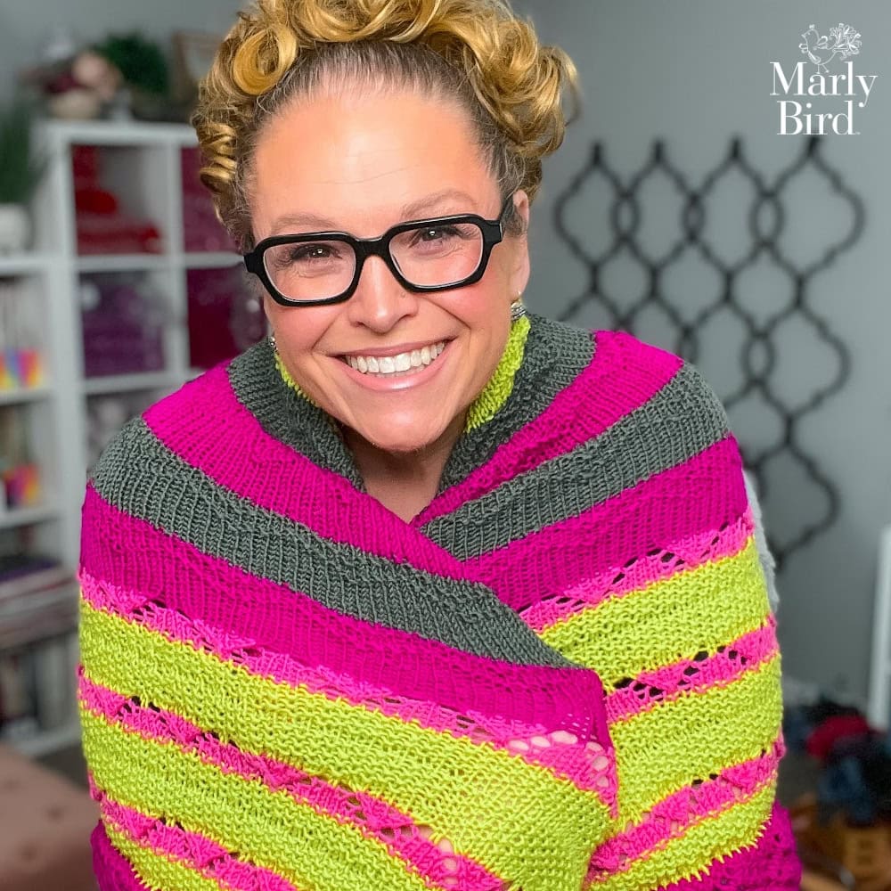 Person wearing a striped pink, gray, and green knit triangle shawl; visible stitch texture; yarn shelves in background.