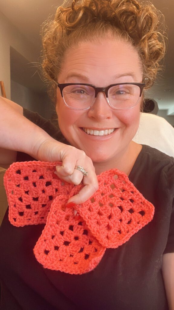 A person wearing glasses smiles while holding up three crocheted Granny Squares in a dimly lit room. They have curly hair and are wearing a black shirt. -Marly Bird