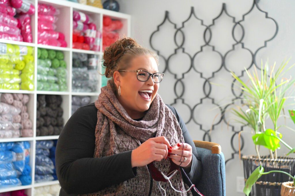 WeCrochet Designer Spotlight on Marly Bird, smiling wearing black and white framed glasses, black tee shirt, and grey knit shawl, sitting in a denim chair and knitting. White ikea shelf full of colorful yarn behind her.
