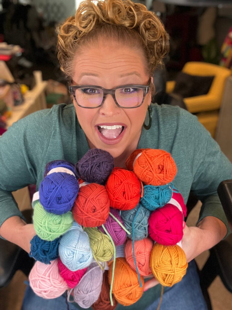 A smiling person with curly hair and glasses holds a large bundle of colorful yarn balls, wearing a green top—ready for the This or That Make-Along 2025—while sitting indoors with blurred background furniture. -Marly Bird