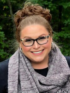 WeCrochet Designer Spotlight - Marly Bird wearing black and white rimmed glasses, black top, and grey knit cowl, with green leafy background.