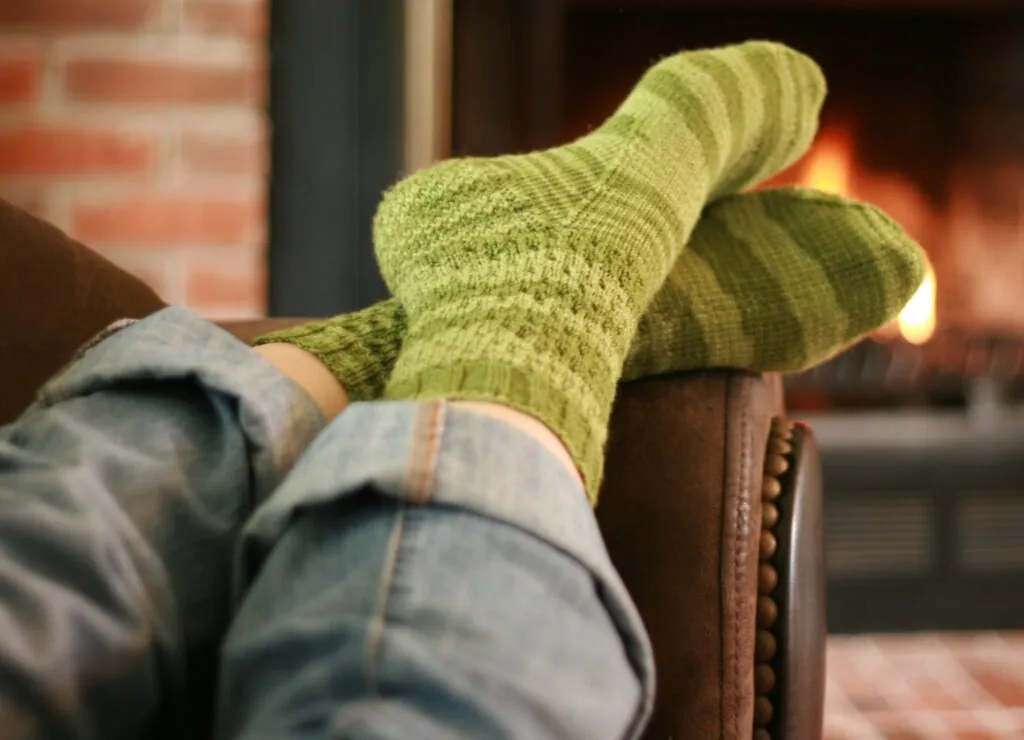 Green hand-knit socks with a cozy ribbed texture, worn with rolled jeans, feet relaxed on a chair by the fireplace.