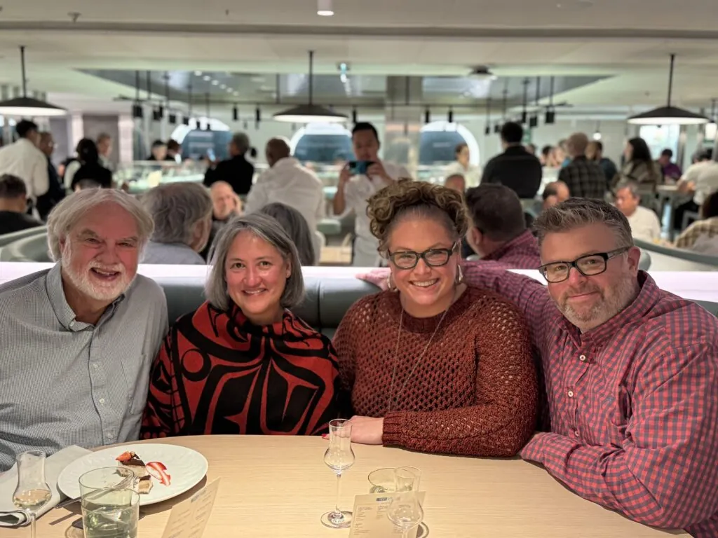 Four adults sit closely together at a well-lit restaurant table, smiling at the camera. One wears a stylish cropped knit sweater. Plates and drinks are in front of them, with other diners filling the busy, modern background. -Marly Bird