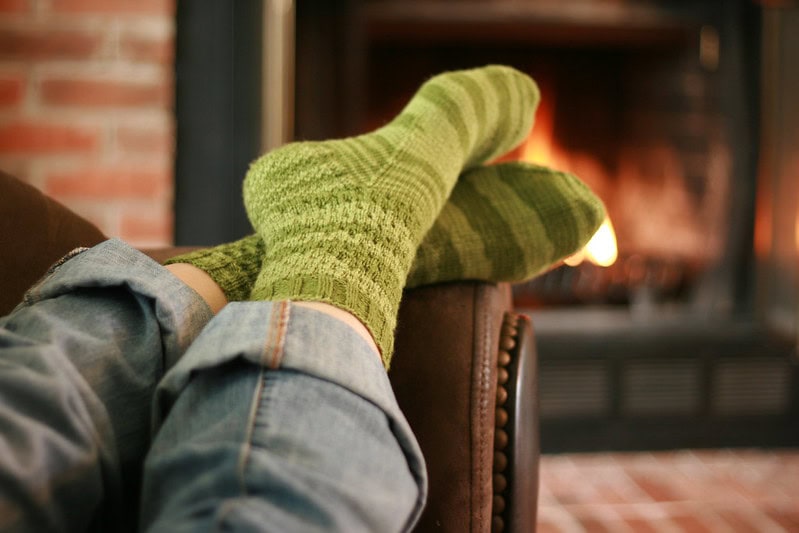 Green crochet socks with visible stitch texture are worn over blue jeans, feet resting on a chair in front of a fireplace.