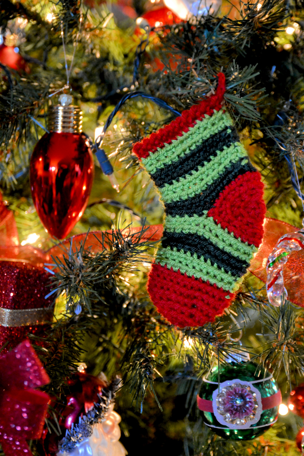 A festive mini crochet Christmas stocking ornament, featuring bold stripes in classic holiday red, green, and black, dangles from a twinkling Christmas tree. The stocking, which exhibits intricate crocheted craftsmanship, is surrounded by an array of traditional decorations including glossy red teardrop baubles, glittery ribbons, and a green ornament with a delicate floral design at its center, all glowing softly amidst the golden fairy lights of the tree. -marly bird