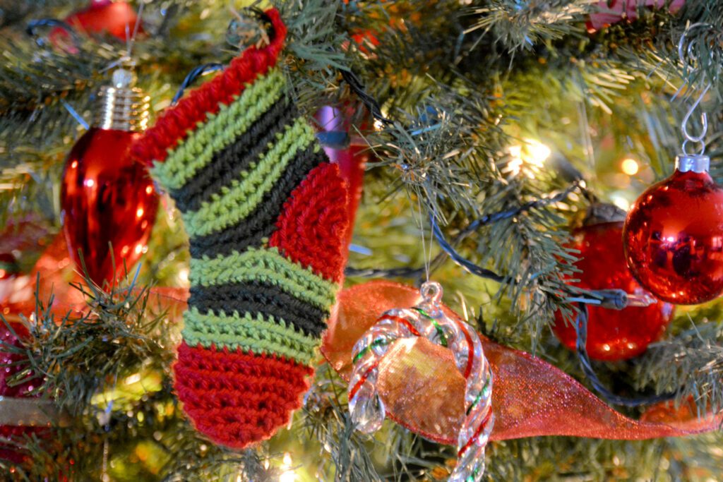 A handcrafted mini crochet Christmas stocking ornament in festive colors of red, green, and black hangs from the branch of a Christmas tree. The stocking features alternating stripes of color, with a ribbed top cuff, and is displayed among twinkling lights and other traditional decorations, including a shiny red ribbon and a translucent red ornament that reflect the warm, soft glow of the holiday lights. - Marly Bird