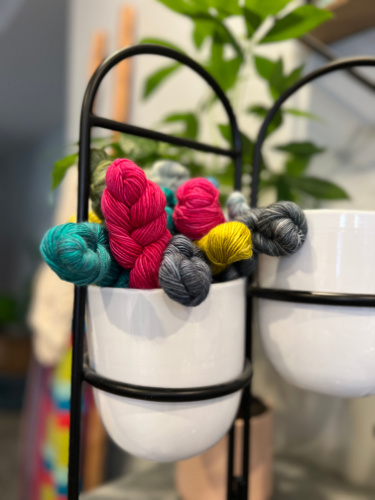 The image shows a collection of mini hanks of yarn artfully arranged in a bowl. The yarns come in a variety of rich colors including teal, pink, grey, yellow, and charcoal, adding a pop of color to the muted backdrop of white bowls. The setting has a creative and cozy atmosphere, indicative of a crafting environment where these yarns might soon be turned into a vibrant knitting project. The image is well composed, with a soft focus that draws attention to the yarns themselves, suggesting they are the main subject, possibly for a crafting or knitting-related theme. -Marly Bird