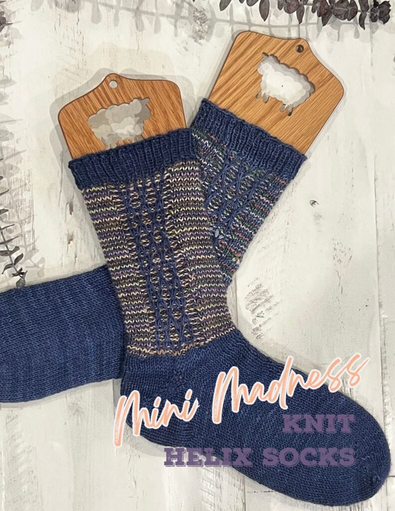 A pair of handmade Marly helix knit socks displayed on wooden sock blockers with the words "mini madness helix knit socks" overlaying the image. The socks feature a colorful striped pattern on a blue base. -Marly Bird