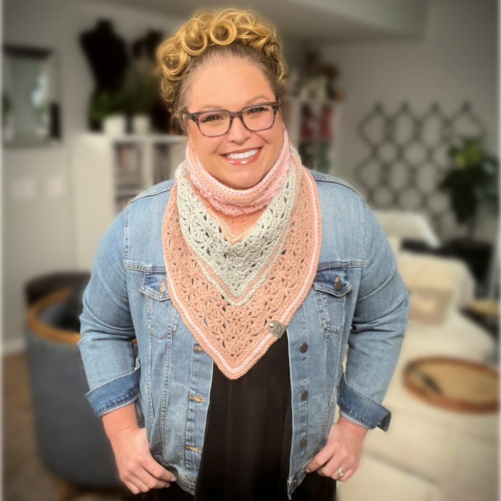 A person wearing glasses smiles warmly, dressed in a denim jacket over a multicolored, crocheted triangular scarf in shades of pink, gray, and cream. The background reveals an indoor setting with plants and blurred decorations. -Marly Bird