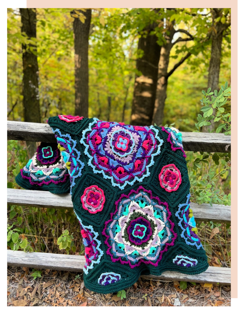 A colorful crocheted blanket with intricate geometric patterns designed by Marly Bird, draped over a rustic wooden fence in a forested area, showcasing vivid shades of pink, green, blue, and black. -Marly Bird