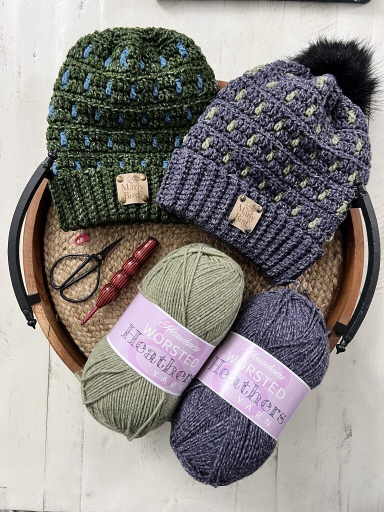 A wooden basket holds two cozy knitted beanies, one green and one gray, each with patterns and labels. Below lie skeins of yarn marked "Worsted Heather." Nearby, crochet hooks suggest the start of colorful crochet mosaic slippers, with scissors ready to complete the creative scene. -Marly Bird