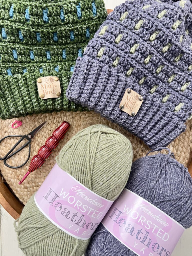 Two crocheted hats in green and blue-gray with "Marly Bird" tags are displayed on a textured surface. Nearby, cozy crochet hooks sit alongside colorful skeins of gray and sage green worsted Heather yarn, ready for your next project. -Marly Bird