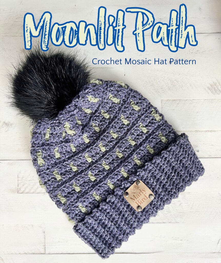 Moonlit Path: Crochet Mosaic Hat Pattern—a cozy crochet hat adorned with a pom-pom, showcasing a vibrant mosaic design in purple and gray tones. A small wooden tag adds charm as it sits on a white wooden surface. -Marly Bird