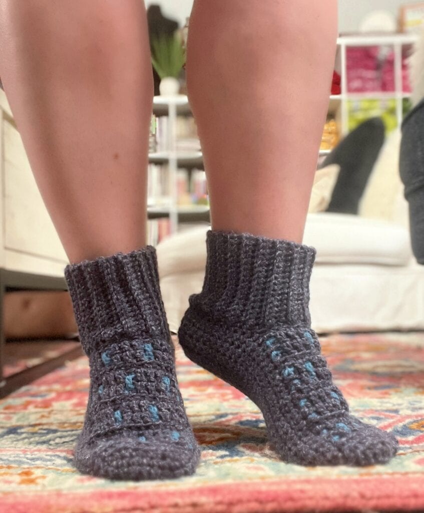 A person wearing dark gray crocheted slipper socks with blue accents stands on a colorful rug, capturing the essence of cozy charm. Only the lower legs are visible, with a white sofa and bookshelf blurred in the background. -Marly Bird