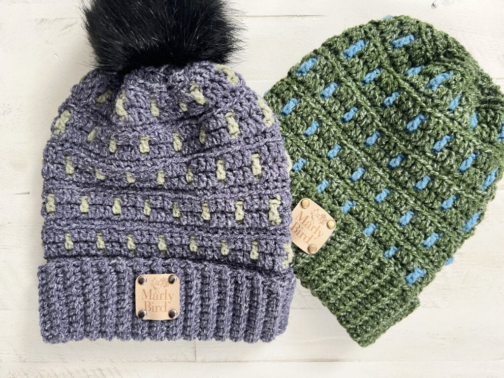 Two crocheted beanies on a white surface: one is purple with light green accents and a black pom-pom, the other is green with blue accents. Both feature a "Marly Bird" tag—perfect inspiration for your own free knitting and crochet patterns. -Marly Bird