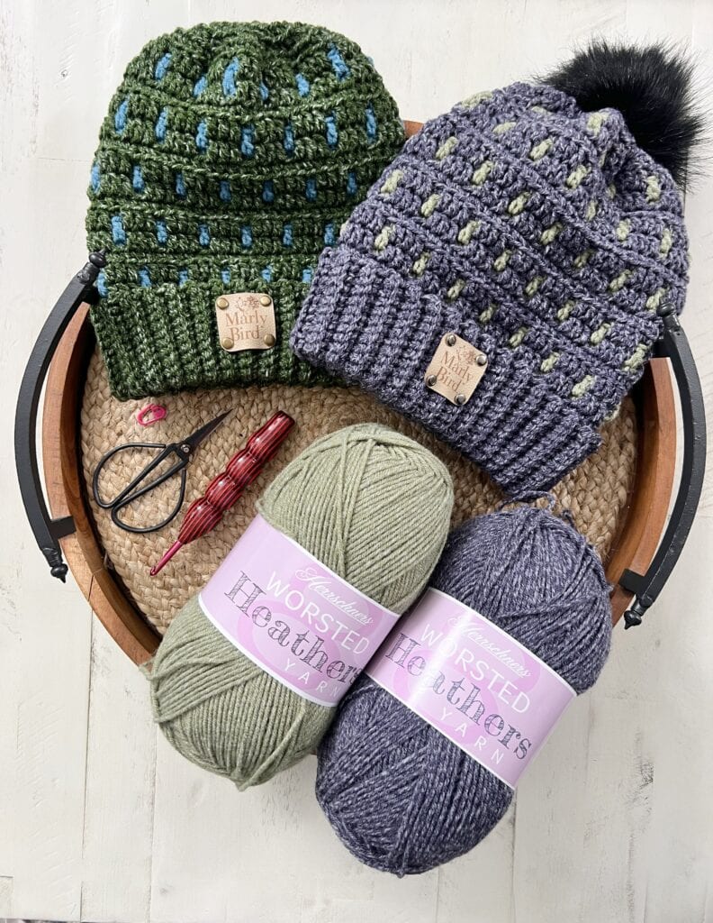 A round basket holds two knitted hats, green and purple, each with a “Mary Bird” tag, plus yarn balls, a pink crochet hook, and small scissors on a woven mat—perfect inspiration for free knitting and crochet patterns. -Marly Bird