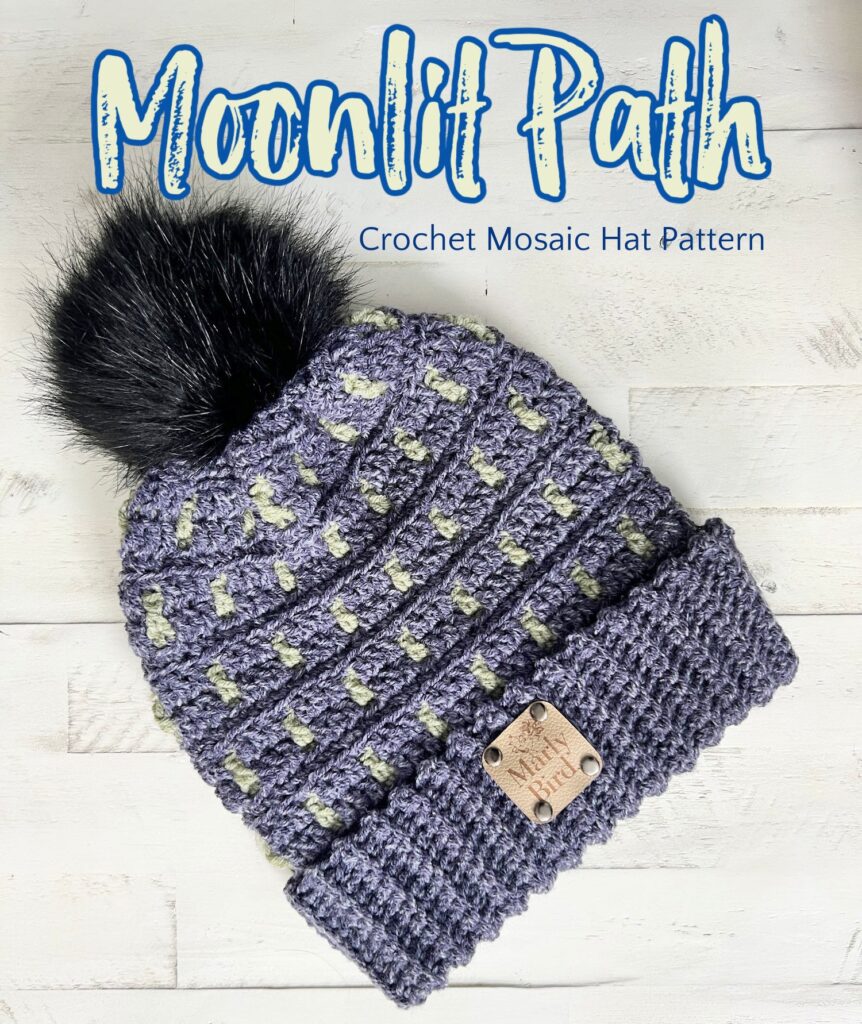 A gray and light green crocheted hat with a black faux fur pom-pom is displayed on a wood surface. The text reads "Moonlit Path Crochet Mosaic Hat Pattern." Discover more free knitting and crochet patterns for stylish accessories. -Marly Bird