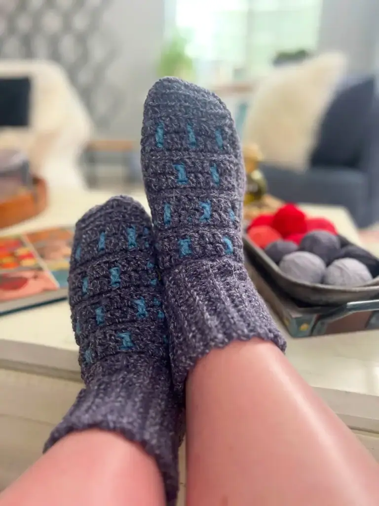 A person’s feet in cozy, hand-knitted gray socks with blue accents rest on a table, alongside balls of yarn, a magazine, and free knitting and crochet patterns in a warm, blurred living room setting. -Marly Bird