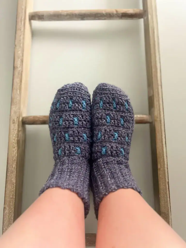 A pair of feet wearing thick, knitted gray socks with blue accents—crafted from free knitting and crochet patterns—rest on a wooden ladder against a white wall. -Marly Bird