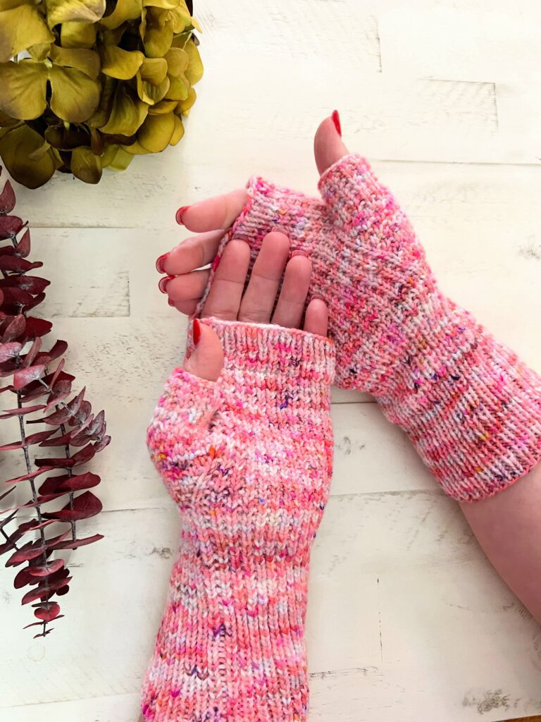 Two hands are adorned with pink speckled knit fingerless gloves. One hand faces up, the other down, revealing the gloves from both sides. Nearby, vibrant dried flowers in green and red rest on a white wooden surface, adding a touch of nature’s charm to this cozy scene. -Marly Bird