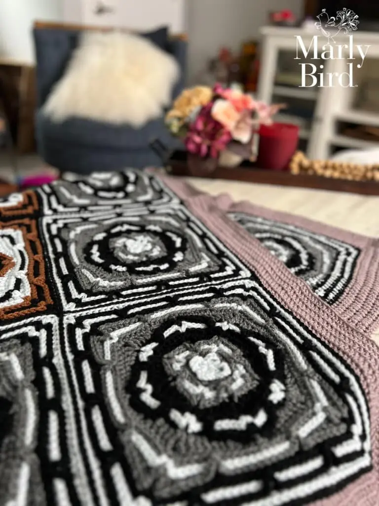 A close-up of a crocheted afghan with intricate black, white, gray, and brown geometric patterns, laid on a table. In the background, a chair, a bouquet, and shelves are visible. "Marly Bird" logo is in the top right corner. -Marly Bird