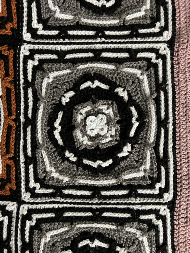 Close-up of a crocheted square pattern with layered geometric shapes in shades of black, grey, and white, bordered by two other crocheted sections in brown and pink. -Marly Bird