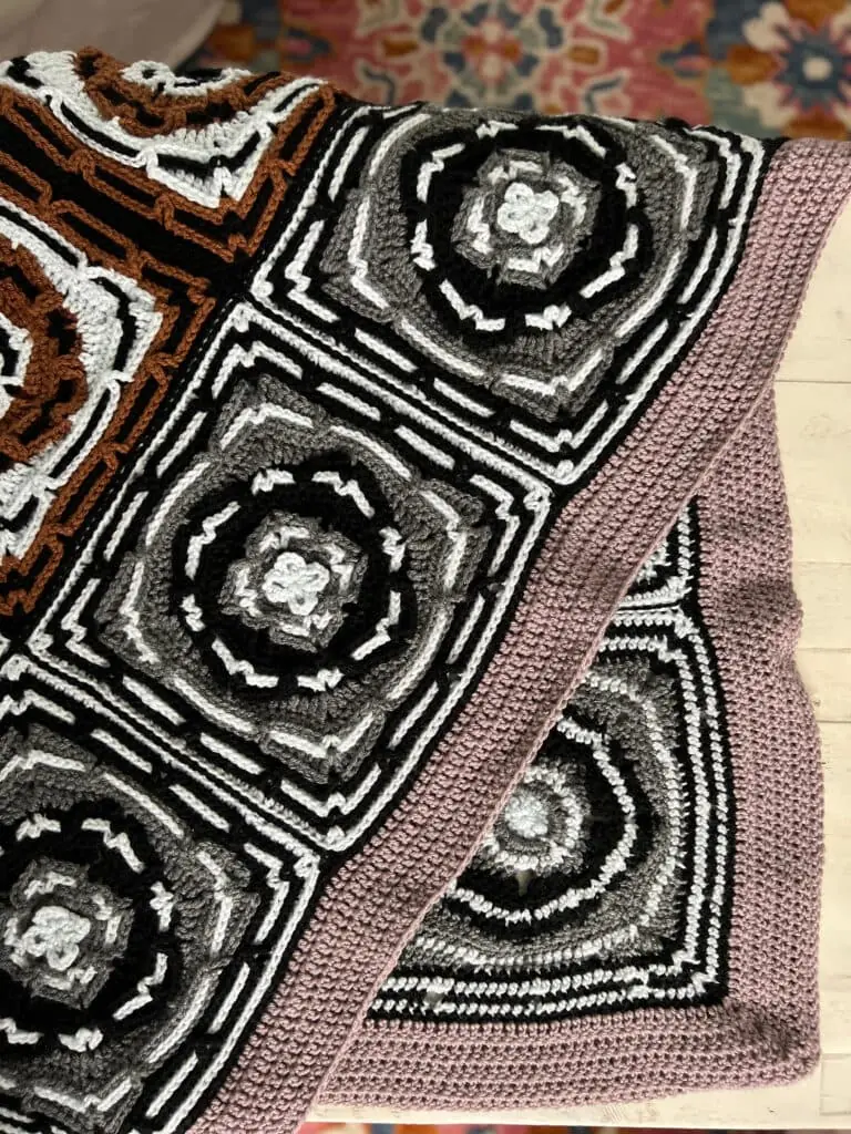 A crocheted blanket with intricate square patterns in black, white, gray, and brown, bordered by a light purple edge. The blanket rests on a light surface with a colorful, floral rug in the background. -Marly Bird