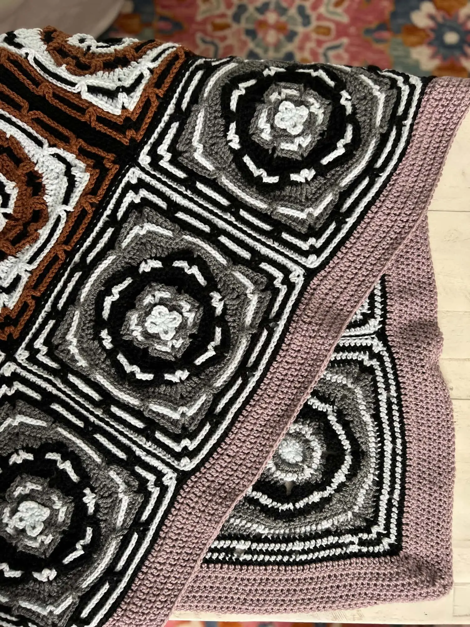 Tunisian crochet blanket with black, white, and gray squares and a dusty pink border, shown on a wood surface and rug.