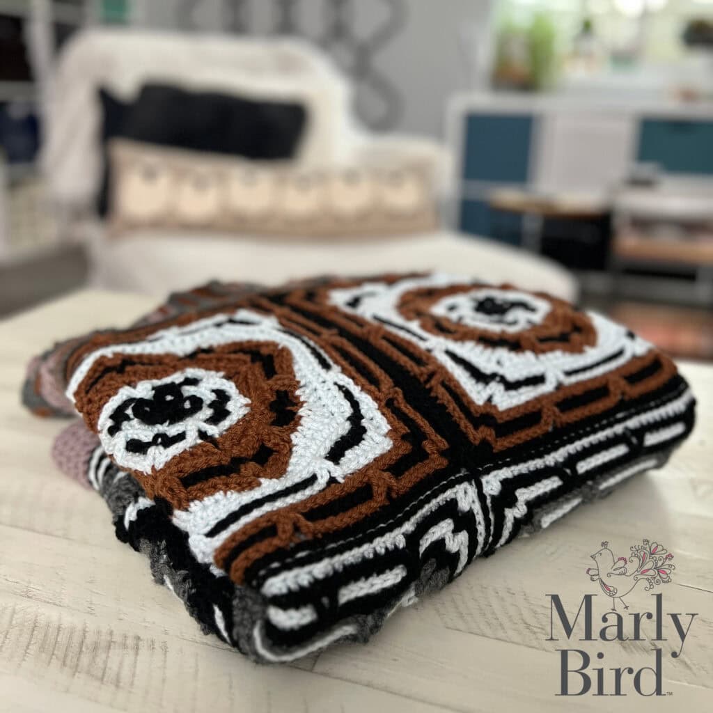 A folded crocheted blanket with geometric patterns in black, white, and brown rests on a wooden table. A blurred living room with a sofa and pillows is in the background. The Marly Bird logo appears in the corner. -Marly Bird