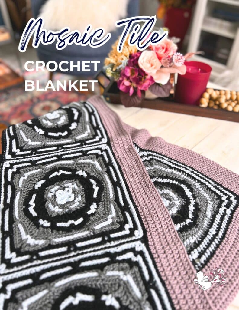 A crochet blanket with a mosaic tile pattern featuring black, white, and gray geometric designs is draped over a beige surface. A bouquet of flowers and decorative items are in the background. The text reads "Mosaic Tile Crochet Blanket. -Marly Bird