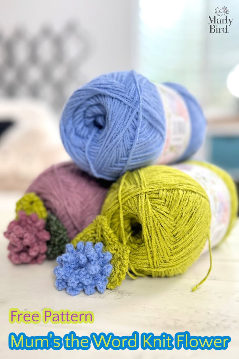 Colorful yarn balls in blue, pink, and green are stacked, adorned with small knitted chrysanthemum flowers. Text reads "Free Pattern: Mum's the Word Knit Flower." Logo in the top right corner against a blurred background. -Marly Bird