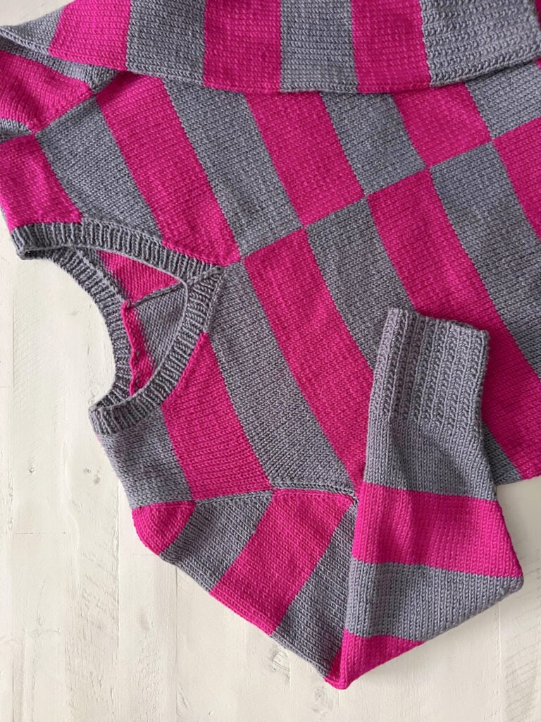 A knit sweater with wide, horizontal stripes in alternating bright pink and gray, laid flat on a white wooden surface. One sleeve is folded across the body of the sweater, showcasing inspiration for free knitting and crochet patterns. -Marly Bird