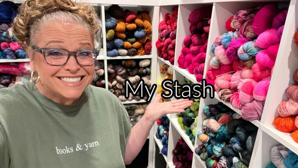 A smiling woman with glasses and curly hair stands beside shelves filled with colorful yarn skeins. She wears a green "books & yarn" shirt and gestures to the yarn with a "My Stash" text overlayed, ready for the This or That Make-Along 2025. -Marly Bird