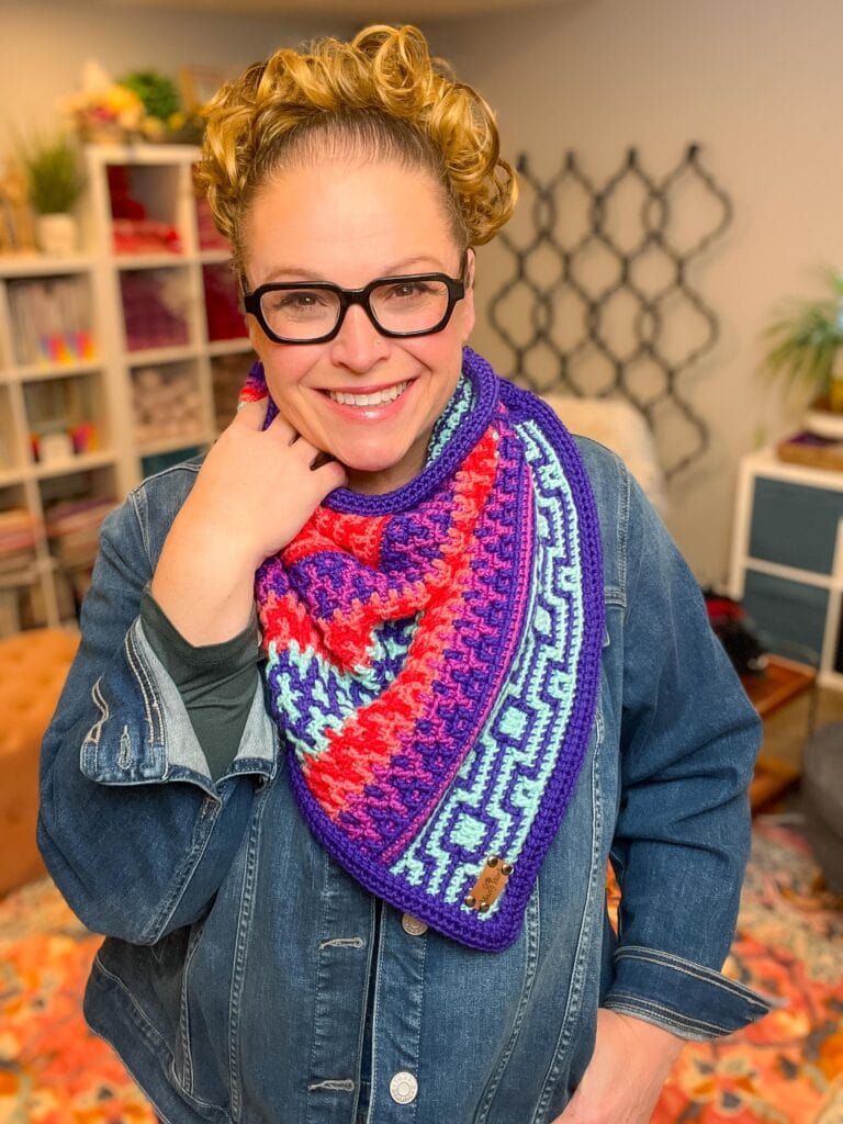 A smiling woman with curly hair and glasses wears a colorful, patterned crochet scarf—perfect for a New Year’s Eve Make-Along—over a denim jacket, standing in a cozy room with shelves and plants in the background. -Marly Bird