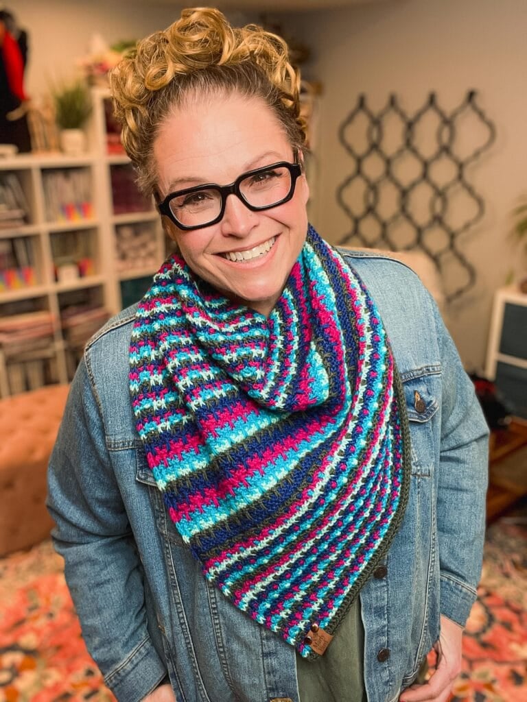 A smiling person with curly hair and glasses wears a blue denim jacket and a colorful, striped crochet scarf reminiscent of box mosaic stitches knitting. Bookshelves and decor are visible in the background. -Marly Bird