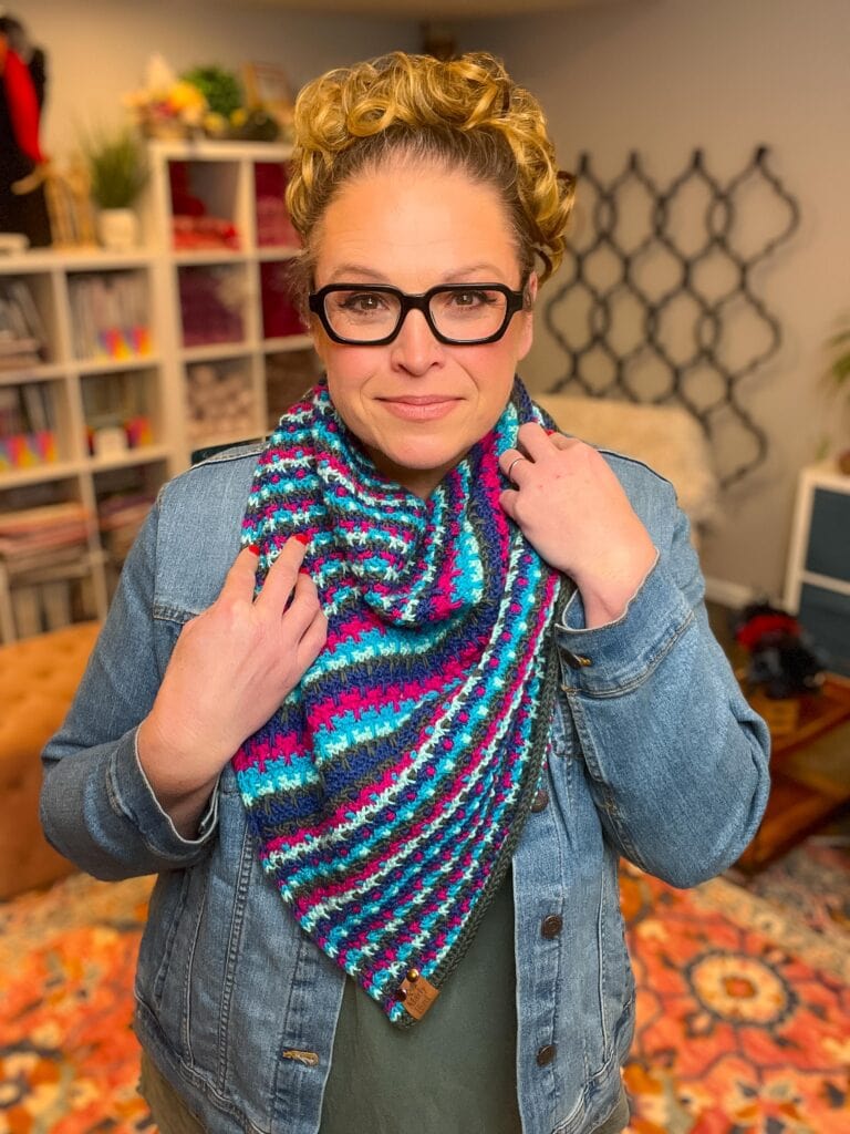 A woman with curly blonde hair and glasses wears a blue denim jacket and a colorful crocheted scarf, perfect for a New Year’s Eve Make-Along. She stands indoors in a cozy room with patterned rugs, shelves, and plants in the background. -Marly Bird