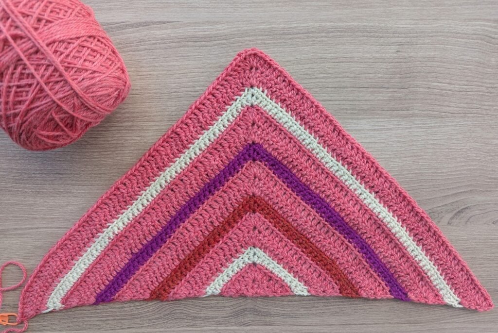 Triangular crochet shawl with pink, red, purple, and cream stripes on wood surface next to a ball of pink yarn.