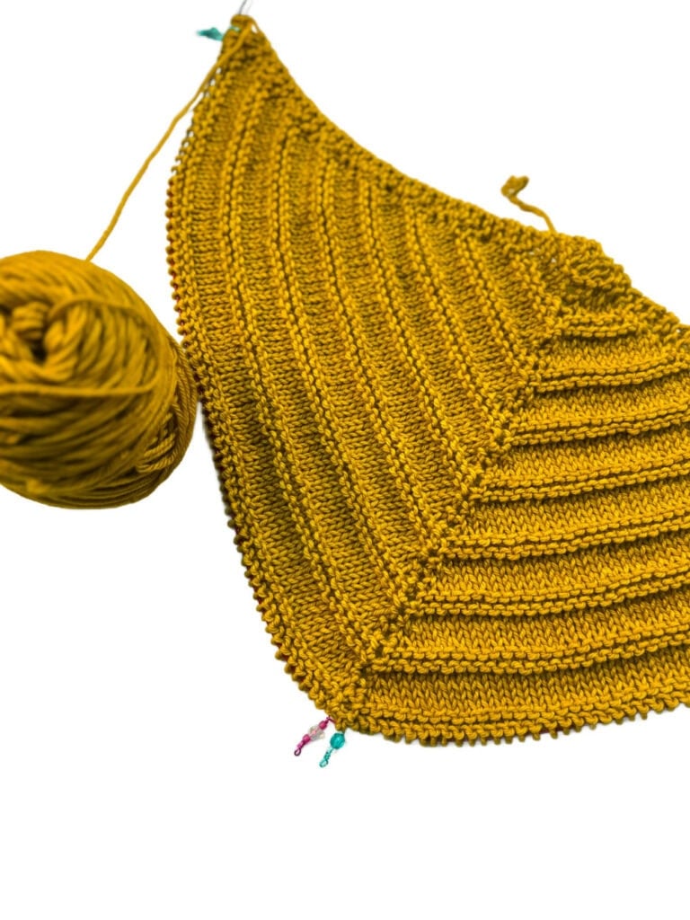 Yellow knitted triangle in progress with matching yarn ball and stitch markers, displayed on a white surface.