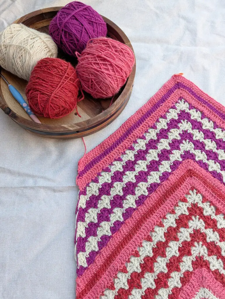 A crochet project with neat colorful stripes, beside a wooden bowl with matching yarn balls and two crochet hooks.