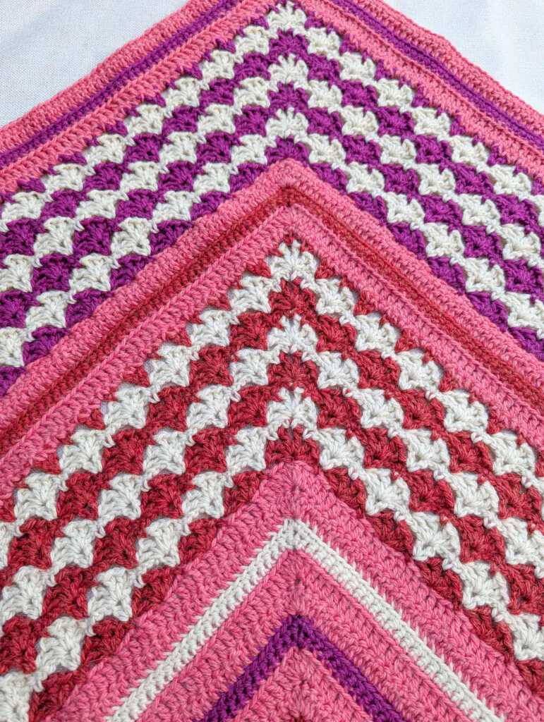 Close-up of a crocheted chevron blanket in pinks, purples, red, and white with textured stitches and detailed patterns.