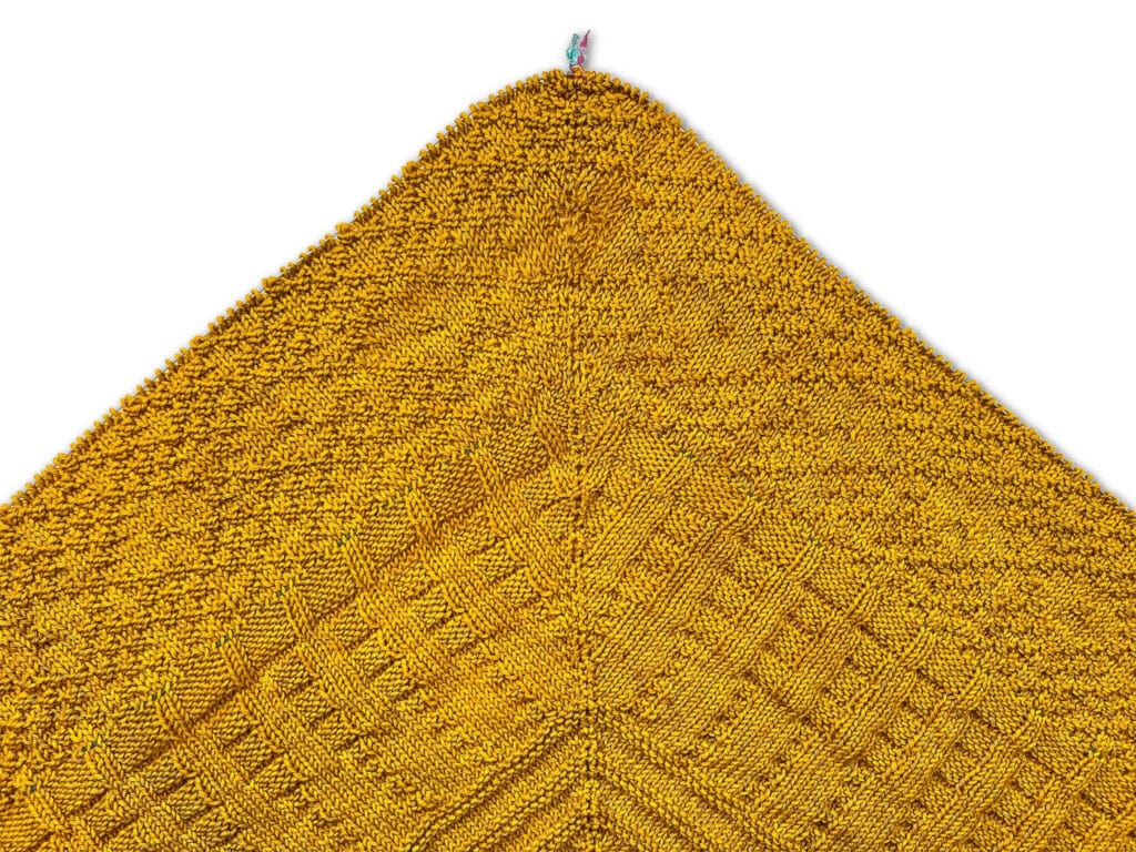 Textured mustard-yellow knit fabric with a diamond pattern and a small colorful stitch marker along the top edge on a white background.