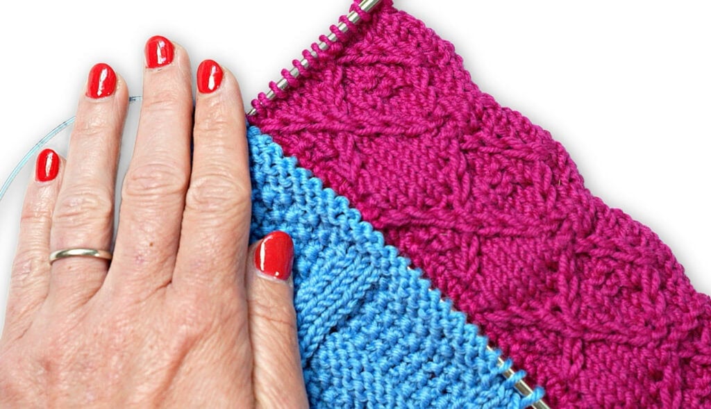 A hand with red nails rests on blue and pink knitting; the pink section shows a diamond textured stitch on needles.