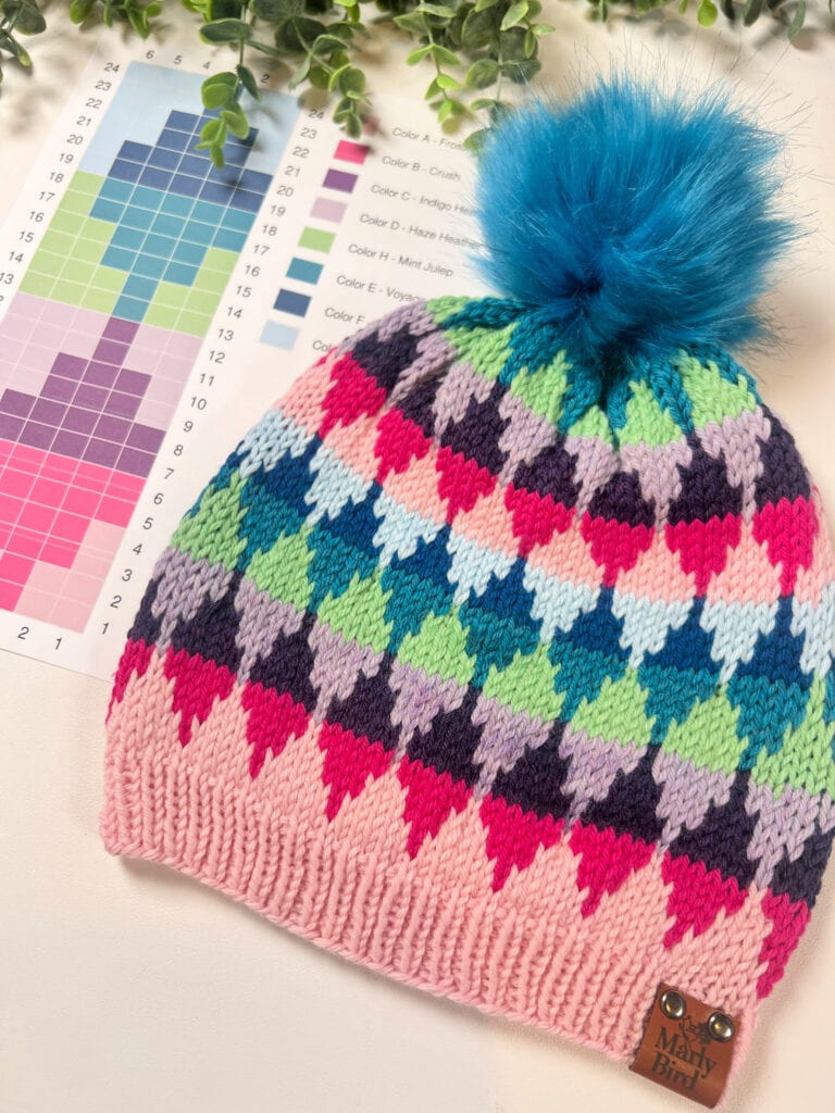 A colorful Knit Hat Pattern with a blue faux fur pom-pom sits on color swatches. The hat features a geometric pattern in pink, blue, purple, and green tones, and a small "Mary Bird" label is attached—perfect for a Northern Gleam vibe. -Marly Bird