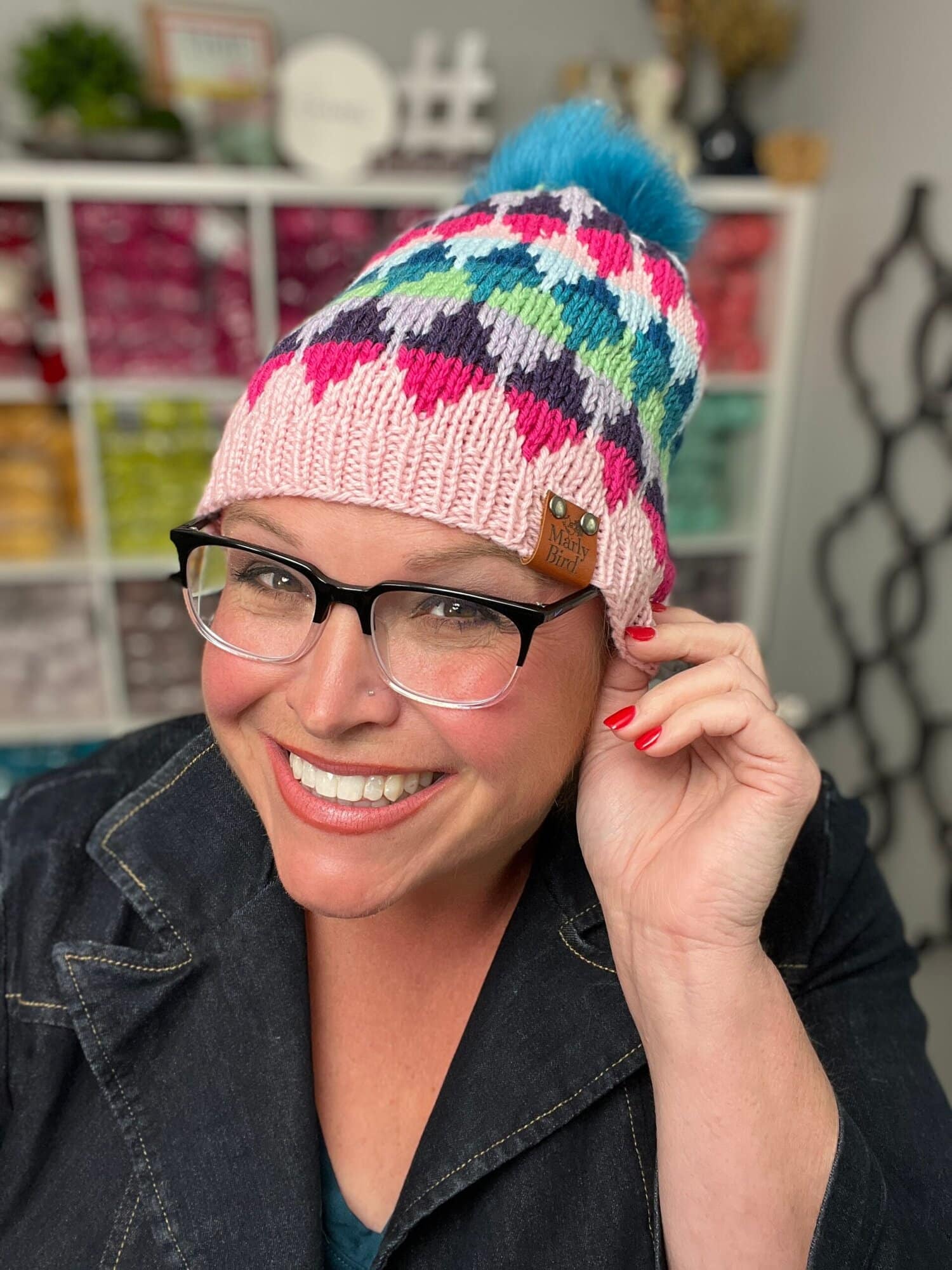 A smiling woman wearing glasses and a colorful knit hat—made from a one-day colorwork knit hat pattern with a blue pom-pom—poses indoors. Shelves of vibrant yarn line the background. She touches her hat’s edge and wears a dark jacket. -Marly Bird