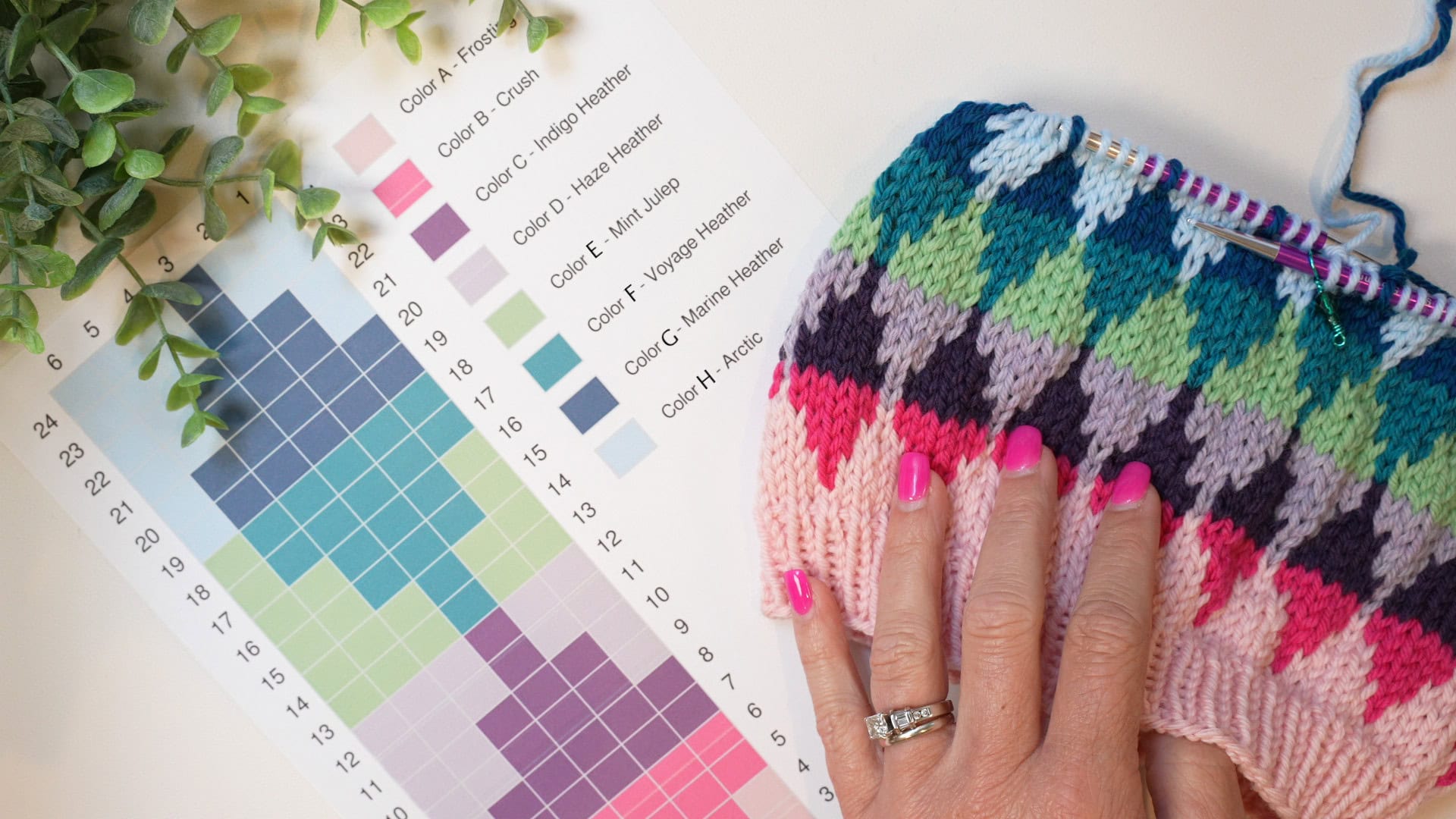 A hand with pink nail polish rests on a colorful knitted piece, showcasing vibrant colorwork next to a color chart with labeled yarn colors and a grid pattern. A green plant is partially visible on the left. -Marly Bird